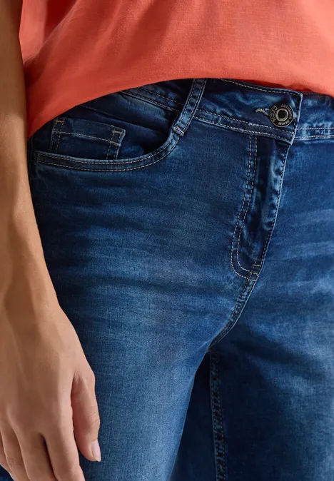 Eine Person trägt ein rotes Shirt und dunkle Jeans, die Hand liegt entspannt neben dem Oberschenkel.